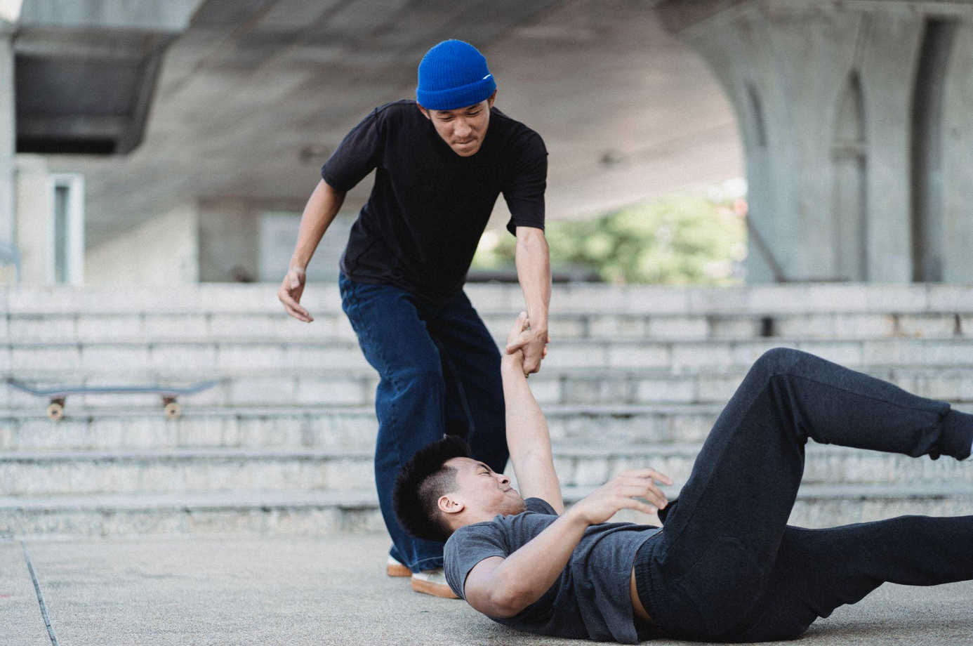 Young Asian man helping friend get up from pavement; image by Allan Mas, via Pexels.com.