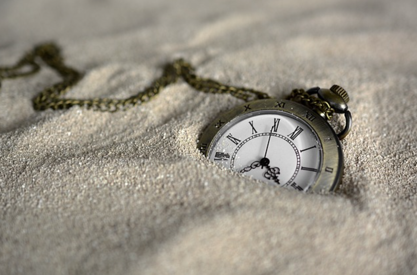 Pocket watch with chain sitting in sand; image by Anncapictures, via Pixabay.com.
