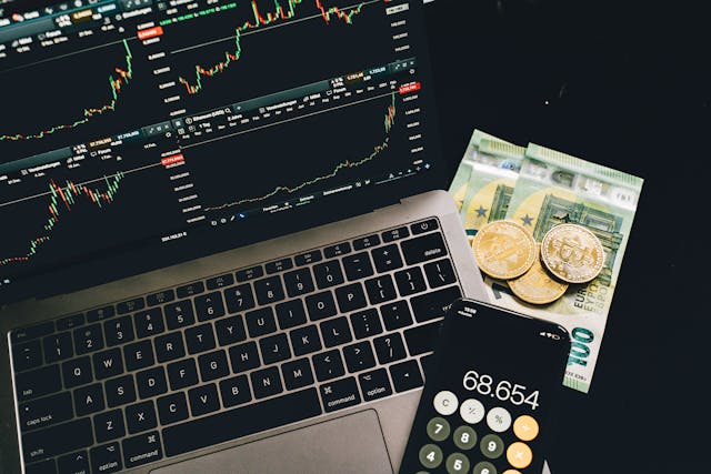 Bitcoins and paper money beside a cellphone and a laptop with graphs on screen; image by Alesia Kozik, via Pexels.com.