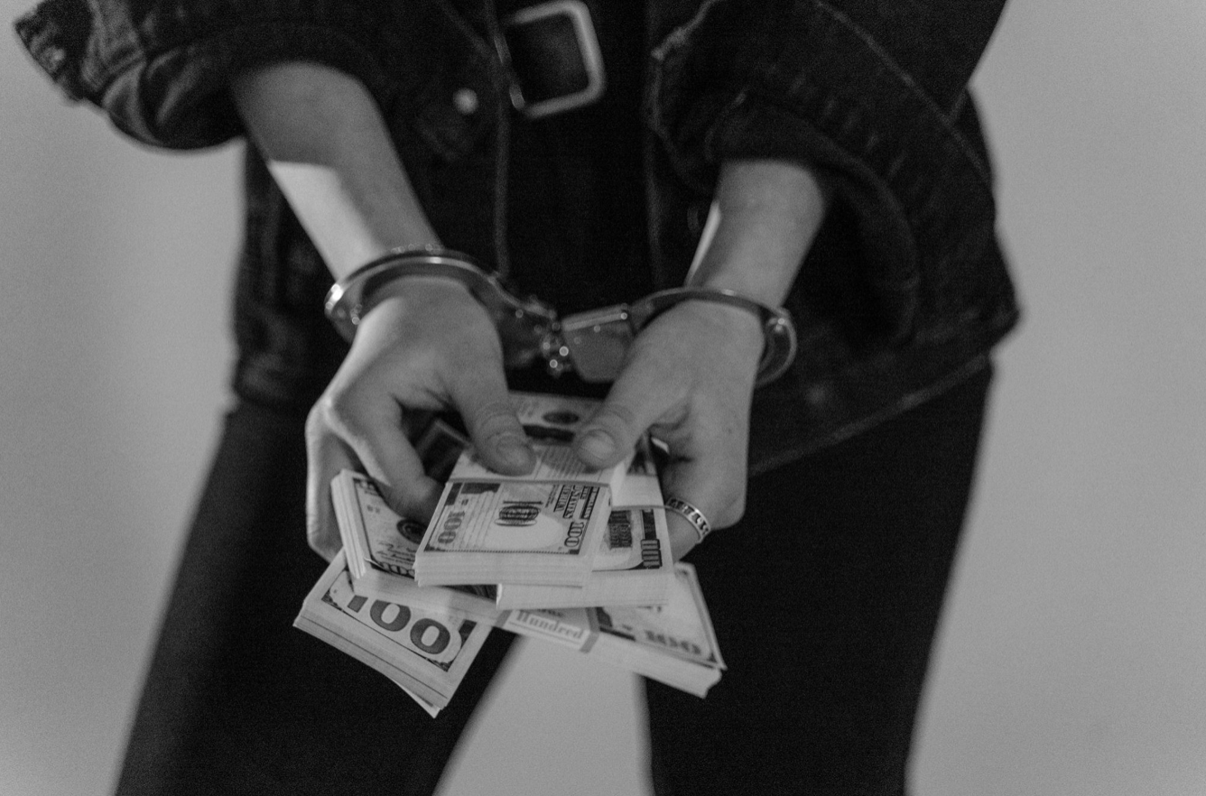 Black and white photo of woman in handcuffs holding five stacks of $100 bills; image by Tima Miroshnichenko, via Pexels.com.