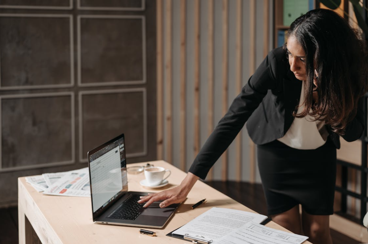 Woman in suit standing at desk working on laptop; image by Pavel Danilyuk, via Pexels.com.