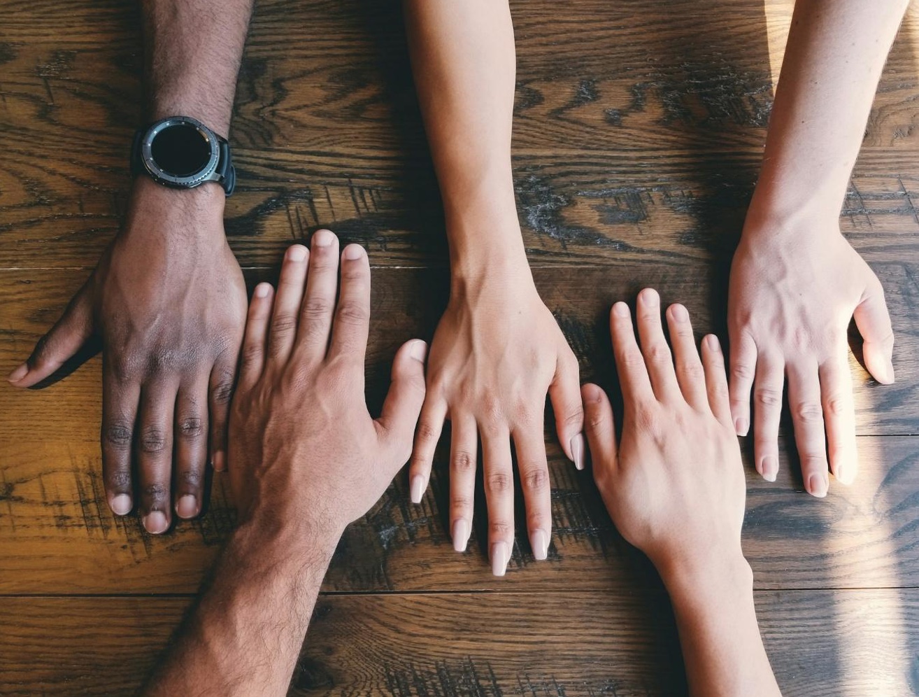 Five human hands on woooden table; image by Clay Banks, via Unsplash.com.