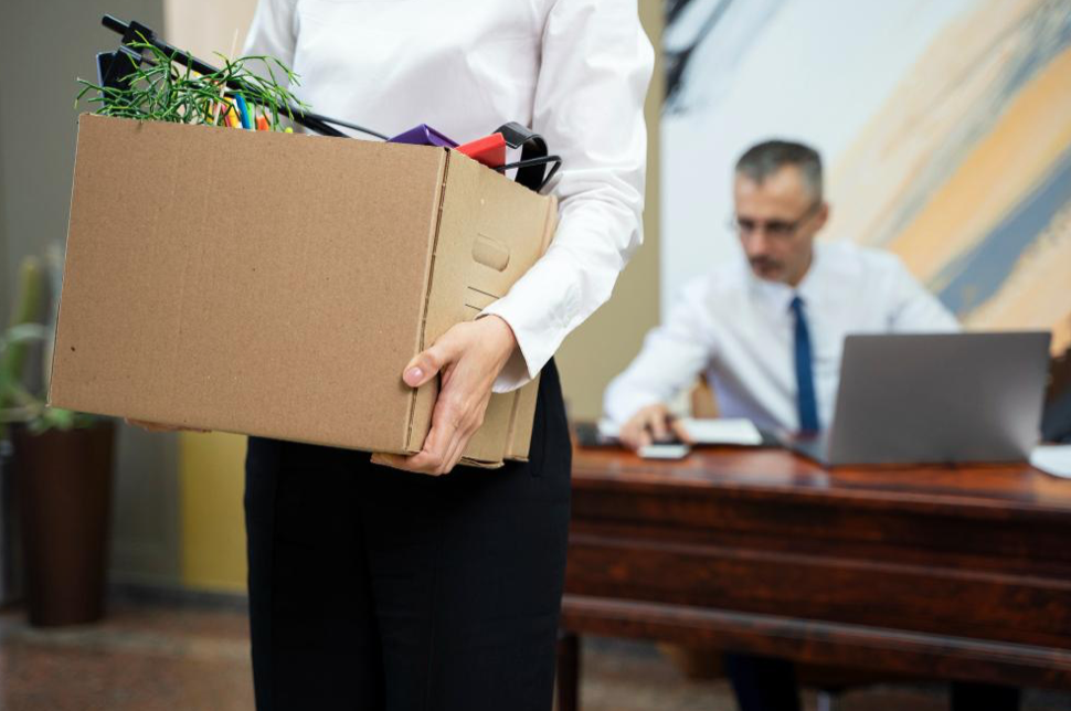 Woman carrying cardboard box full of personal items from her office; image by Freepik, via Freepik.com.
