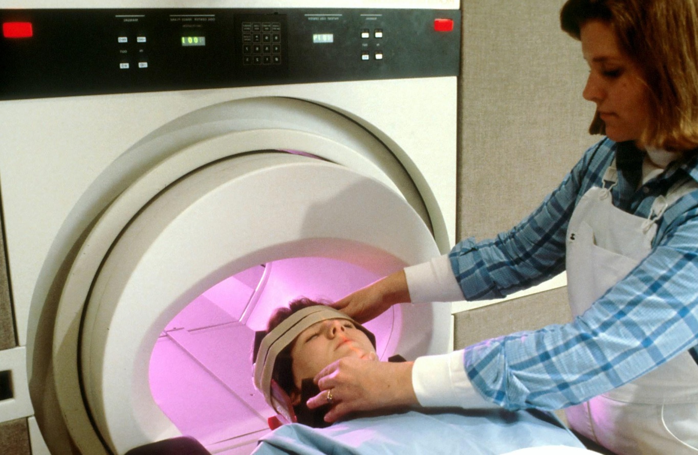 A Caucasian woman's head is being secured by a Caucasian female technician, preparing the patient for magnetic resonance imaging (MRI). Image by National Cancer Institute, via Unsplash.com.