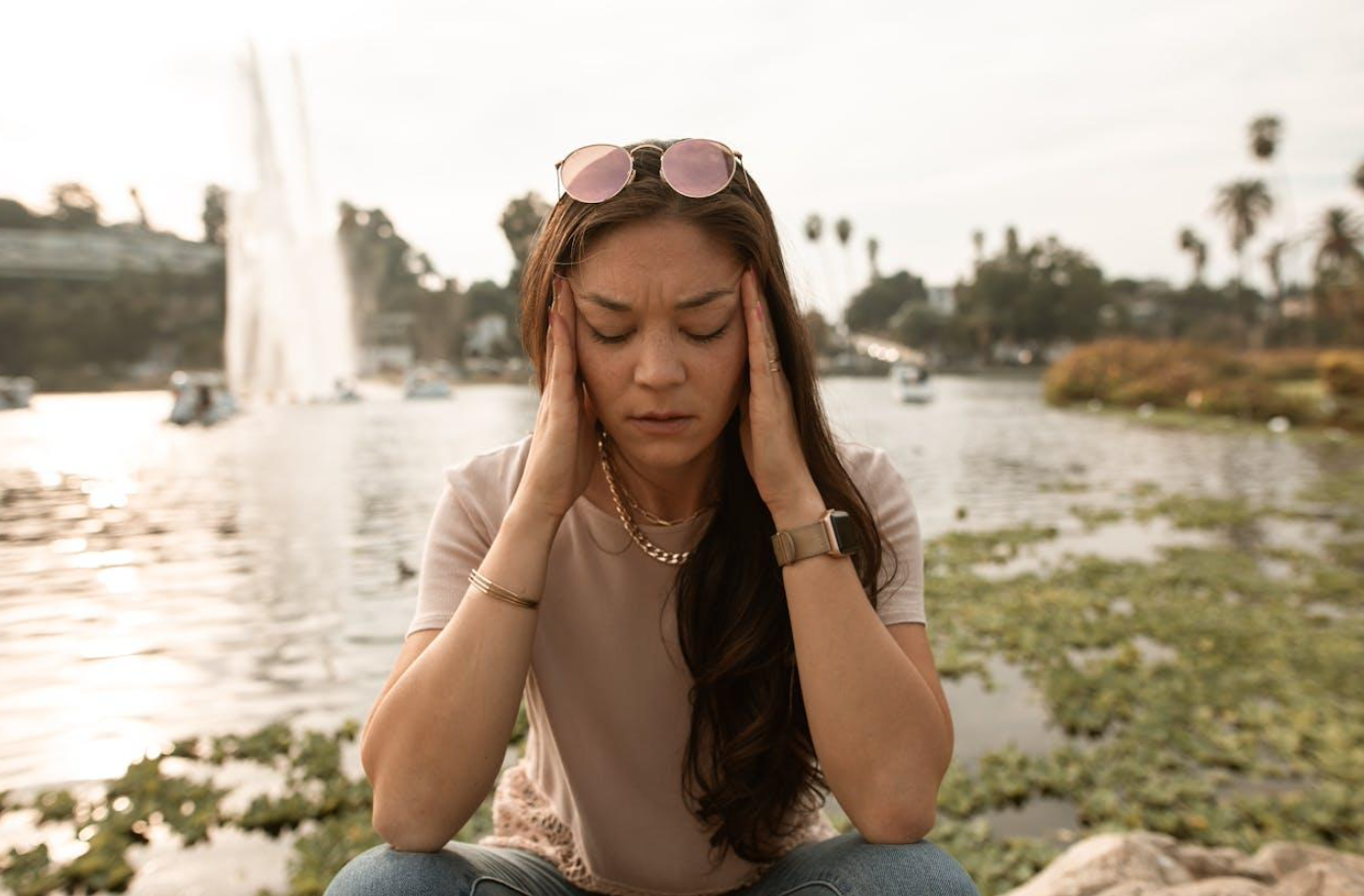 Stressed out woman sitting by lake massaging her temples; image by RDNE Stock project, via Pexels.com.