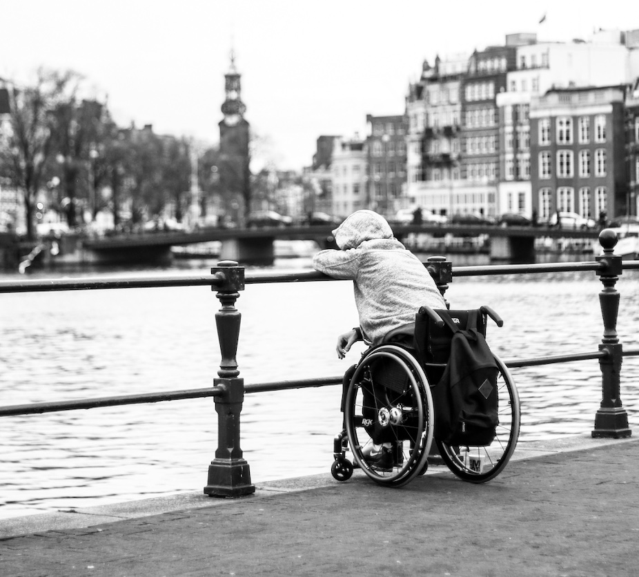 Grayscale photo of main in wheelchair on bridge; image by Ben Allan, via Unsplash. Cropped for use.
