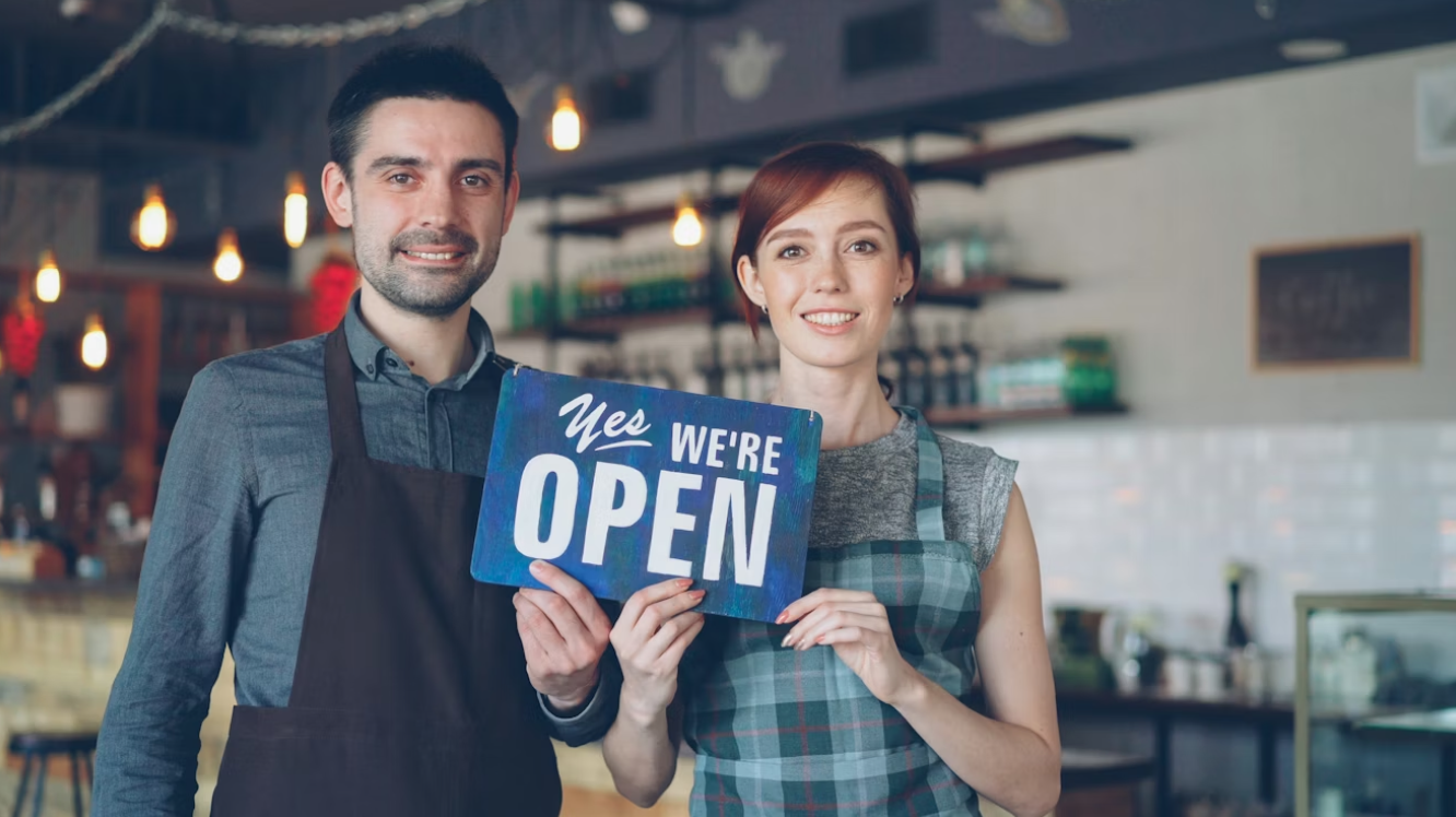 Smiling couple holds open sign; image by Vitaly Gariev, via Unsplash.com.