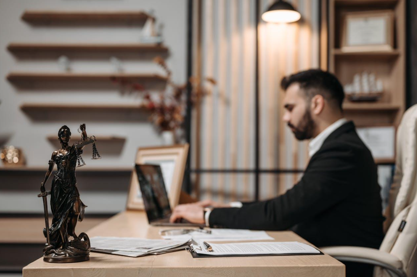 Soft focus shot of lawyer at desk with sharp focus statue of Lady Justice in the foreground; image by Pavel Danilyuk, via Pexels.com.
