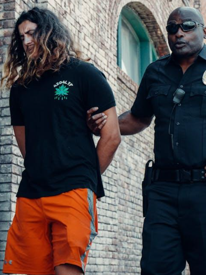 Police officer escorting handcuffed man with long hair near brick wall; image by Kindel Media, via Pexels.com.