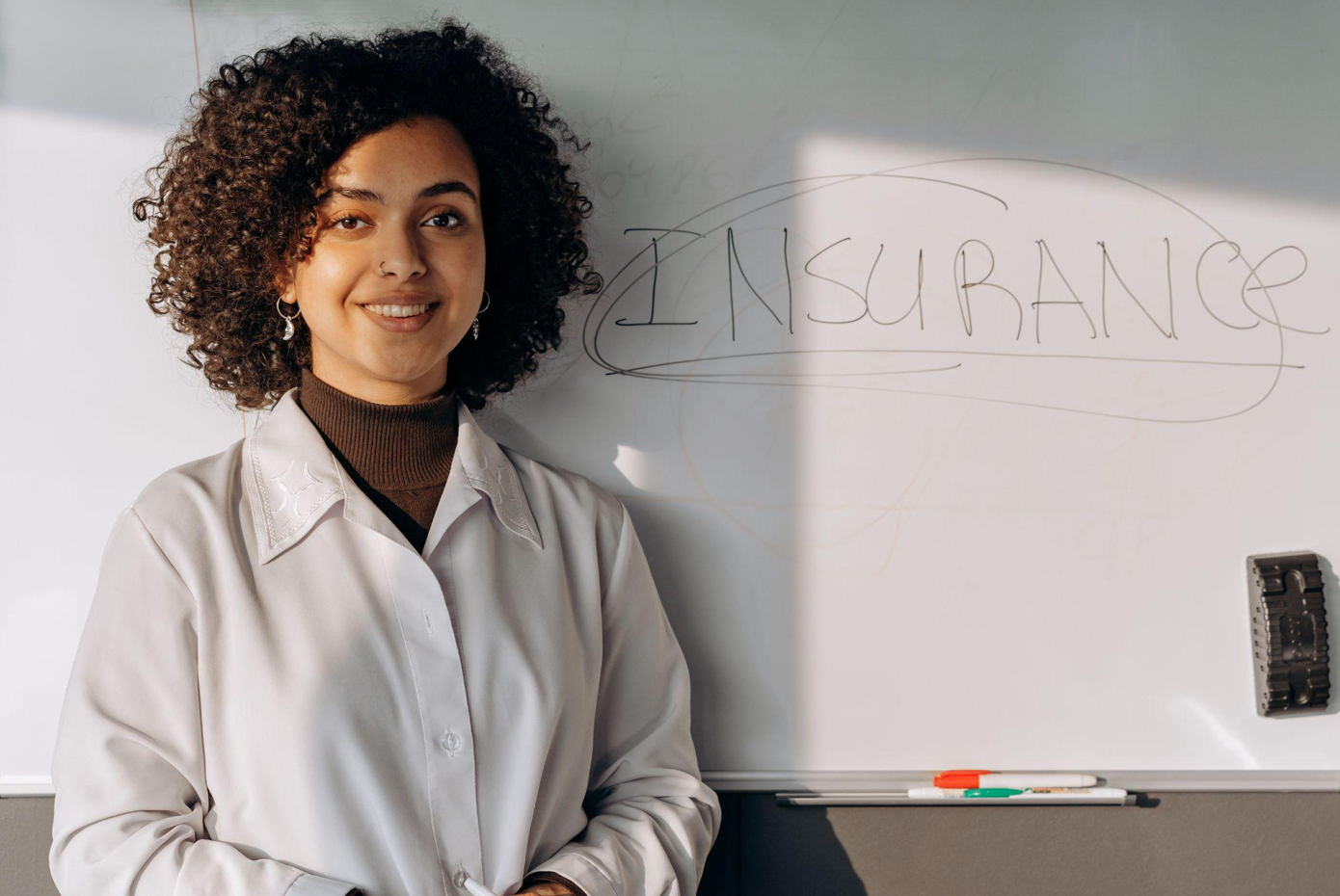 African American woman smiling in office setting with a whiteboard displaying 'Insurance'; image by Mikhail Nilov, via Pexels.com.