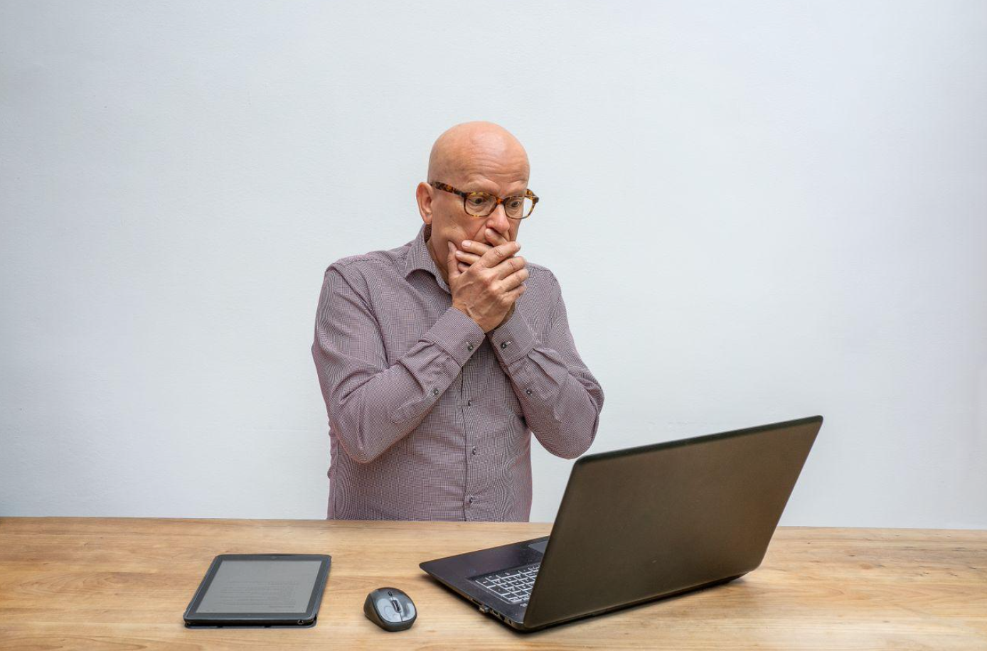 Man in gray longsleeve shirt with hands over his mouth in surprise, looking at laptop screen; image by Mike van Schoonderwalt, via Pexels.com.