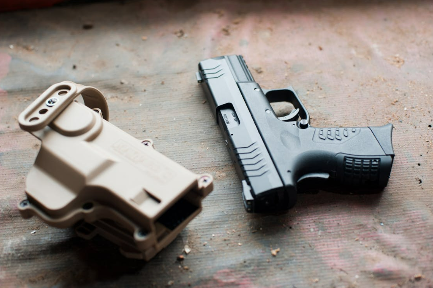 Black pistol beside beige gun case on dirty surface; image by Dan Galvani Sommavilla, via Pexels.com.